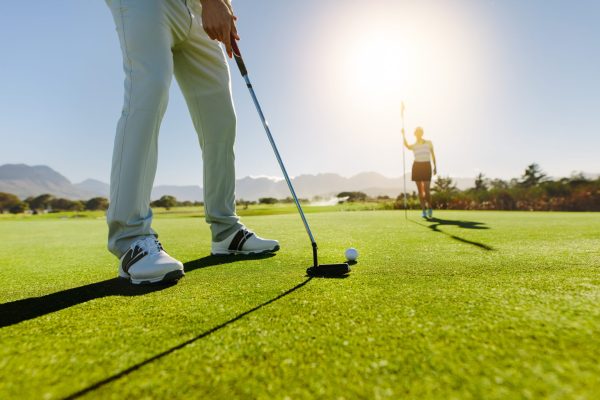 Low angle view of golfer on putting green about to take the shot. Male golf player putting on green with second female player in the background holding the flag.